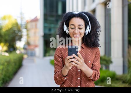 Eine lächelnde junge Frau mit lockigem Haar hört Musik über Kopfhörer, während sie ihr Telefon hält. Sie scheint zufrieden und entspannt zu sein. Stockfoto