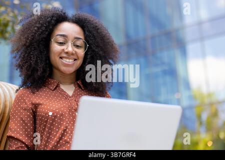 Eine junge Frau mit Brille arbeitet draußen an einem Laptop und lächelt glücklich. Das natürliche Licht verstärkt ihr angenehmes Verhalten. Stockfoto