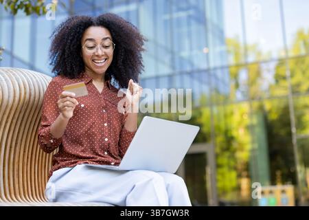 Eine freudige Frau kauft online mit einem Laptop und einer Kreditkarte im Freien ein und genießt dabei den Sonnenschein und die Stadtlandschaft. Stockfoto