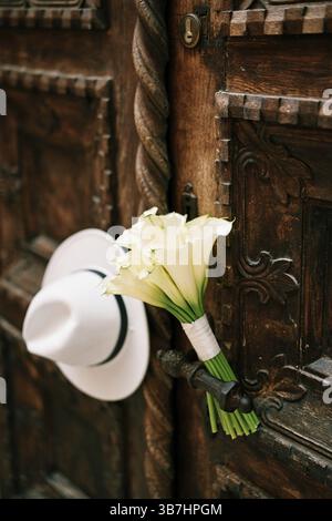 Ein Brautstrauß und ein weißer Hut hängen am Metallgriff einer antiken Holztür. Stockfoto