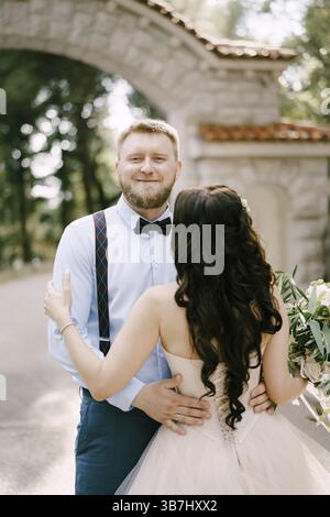 Die Braut umarmt den Bräutigam im Park neben einem wunderschönen Backsteinbogen, Rückblick. Stockfoto