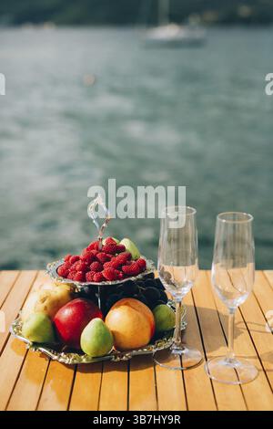 Zweistöckiger Teller mit Früchten: Himbeeren, Pfirsich, Äpfel, Feigen, Birnen, Trauben. Und zwei Gläser Champagner auf dem Tisch am Meer Stockfoto