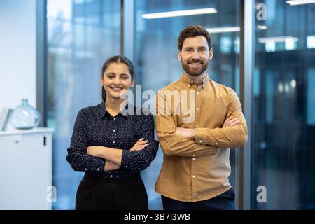 Lächelnde Geschäftsleute stehen mit gekreuzten Armen in einer hellen, modernen Büroumgebung zusammen und sind bereit für den Erfolg. Stockfoto