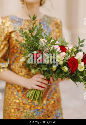 Eine Braut in einem ungewöhnlichen goldenen Kleid hält einen Brautstrauß aus weißen und roten Rosen in der Hand, Nahaufnahme. Stockfoto