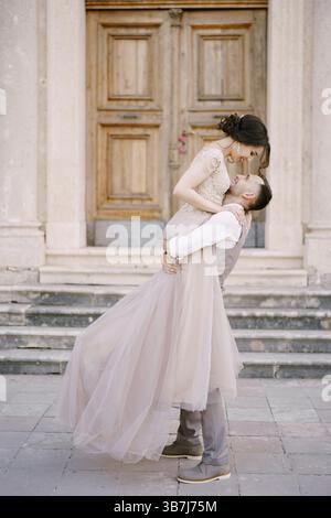 Der Bräutigam hält die Braut in den Armen vor dem Eingang des alten Gebäudes. Stockfoto