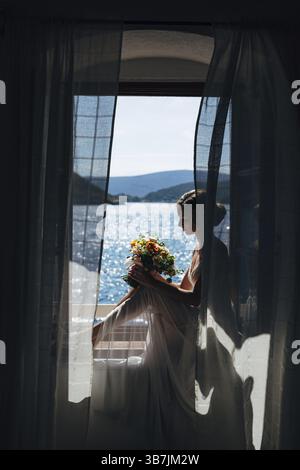 Die Braut sitzt an einem offenen Fenster mit Meerblick und flatternden Vorhängen und hält einen Hochzeitsstrauß mit orangefarbenen Blumen. Stockfoto