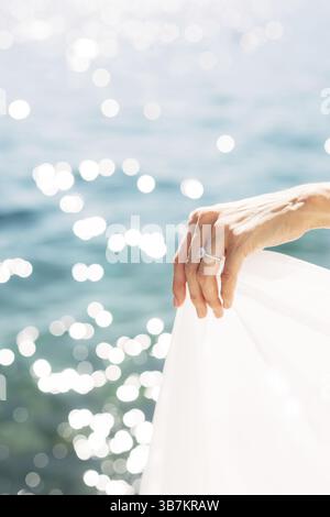Braut Hand mit einem Ehering am Finger, Nahaufnahme. Die Braut hält den Rand des Kleides vor dem Hintergrund des Meeres. . Stockfoto