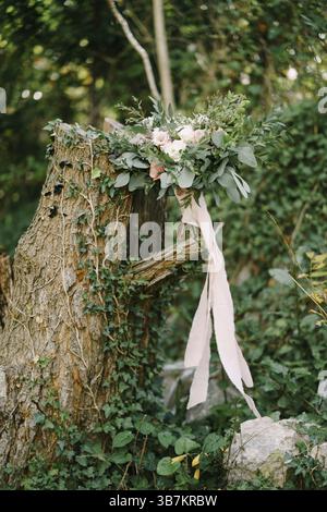 Brautstrauß aus weißen und rosa Rosen, Eustoma, Zweigen von Eukalyptusbaum und Buchsbaum, astilbe und rosa Bändern auf einem trockenen, mit Efeu bedeckten Stumpf. Stockfoto