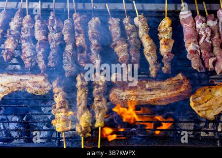 Ein Grill gefüllt mit Spießen und Schweinebauch, der über offenen Flammen kocht. Rauch steigt, wenn das Fleisch bräunlich wird, die Szene zeigt ein traditionelles Barbecue in Stockfoto