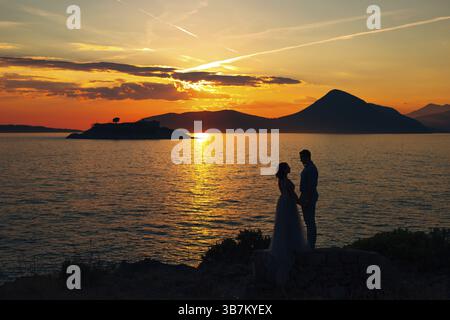 Die Braut und der Bräutigam stehen am Strand der Insel Mamula und halten Hände am Sonnenuntergang. Stockfoto
