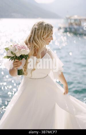 Eine Braut in einem zarten Kleid mit einem Blumenstrauß in der Hand steht auf einem Pier in der Bucht von Kotor . Stockfoto
