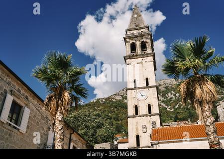 Glockenturm der Kirche St. Nikolaus mit einer Uhr zwischen alten Häusern vor dem Hintergrund grüner Berge. Perast, Montenegro. Stockfoto