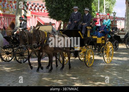 Traditionelle Pferdekutsche auf der Feria de Sevilla 2025 mit Reitern in formeller Kleidung und Frauen in farbenfroher Flamenco-Kleidung, die die festliche Parade genießen. Stockfoto