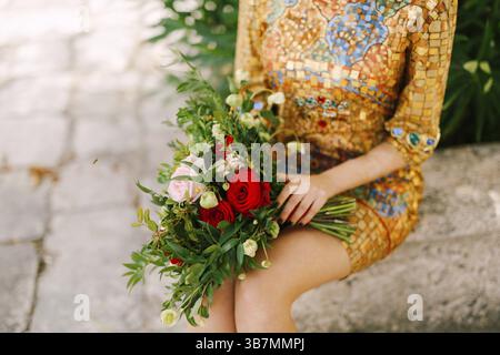 Eine Braut in einem ungewöhnlichen goldenen Kleid mit Ikone sitzt und hält einen Brautstrauß aus weißen und roten Rosen in der Hand, Nahaufnahme. Stockfoto