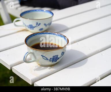 Zwei wunderschön gestaltete Tassen mit dunklem Kaffee stehen auf einem weißen Holztisch in einem hellen Garten. Das Sonnenlicht verstärkt den Moment und schafft eine Ruhe Stockfoto