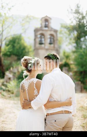 Die Braut und der Bräutigam in Kränzen umarmen sich am alten Glockenturm in der Nähe der Kirche in Prcanj. Stockfoto