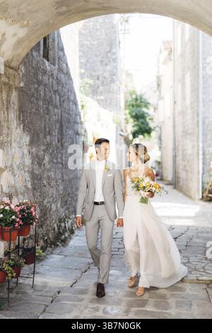 Die Braut und der Bräutigam mit einem Blumenstrauß gehen Hand in Hand entlang einer gemütlichen Straße der Altstadt unter dem Bogen und lächeln. Stockfoto