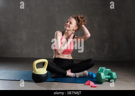Junge Frau in Sportbekleidung, die auf Yogamatte sitzt, Nackenschmerzen nach dem Training spürt, Muskelschmerzen berührt. Stockfoto
