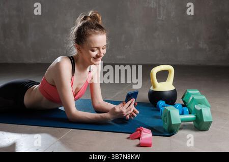 Lächelnde Frau, die sich nach dem Workout entspannt, auf der Matte liegend und im Fitnessstudio mit Wasserkocher und Hanteln mit Smartphone benutzt. Stockfoto