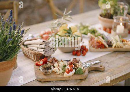Holzschneidebrett mit Köstlichkeiten und eine Schale Oliven auf dem Tisch mit Zitronen, eine Blume in einem Topf und ein Korb mit Servietten. Stockfoto