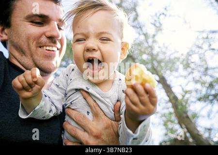 Der lächelnde Vater hält ein lachendes Kind mit einer Birne in der Hand vor einem Hintergrund aus Holz. Nahaufnahme. Niedriger Winkel. Stockfoto