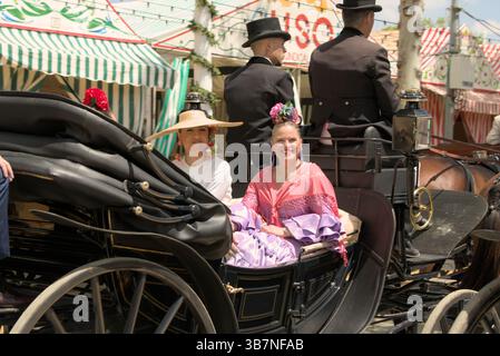 Lächelnde Frauen in Flamenco-Kleidern fahren während der Feria de Sevilla 2025 in einer traditionellen Pferdekutsche. Feria de Sevilla, Spanien Stockfoto