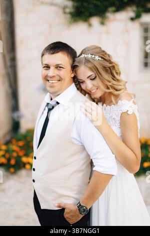 Die Braut umarmt den Bräutigam auf den Straßen von Perast neben weißen Ziegelhäusern. Stockfoto