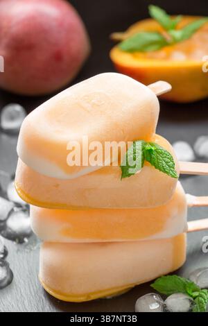Spalte mit 4 Mangosorbet Eisbecher mischen Eis hinter Mango und geschnittener Mango in der Nähe von Eiswürfeln auf schwarzem Hintergrund. Mangosorbet Eisbecher mischen Eis Eis Eis Eis Eis Eis Eis Eis. Vertikal. Stockfoto