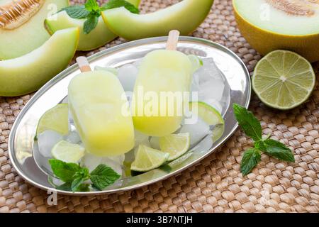 Melonen- und Limettensorbet-Eis auf Silberplatte mit Eiswürfeln, Limettenstückchen, Minzblätter in der Nähe von Melonenscheiben, Limette auf einem Strohserviettenhintergrund. Melonen- und Limettensorbet-Eis-Eis. Horizontal. Stockfoto