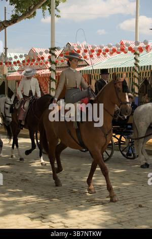 Andalusierin auf einem Pferdezug in La Feria de Sevilla 2025, umgeben von farbenfrohen Kasetten und Laternen. Feria de Sevilla, Spanien Stockfoto
