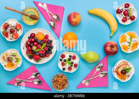 6 Schüsseln Joghurt mit verschiedenen Zutaten Obst, Beeren, Nüssen, Honig und 6 Löffeln, Servietten und ein paar leere Stellen auf hellblauem Hintergrund. Horizontal. Draufsicht. Stockfoto