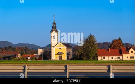 Domzale, Slowenien - 29. Oktober 2024: Malerische ländliche Szene mit einer gelben Kirche mit einem hohen Turm, umgeben von Häusern und Bäumen, Set ag Stockfoto