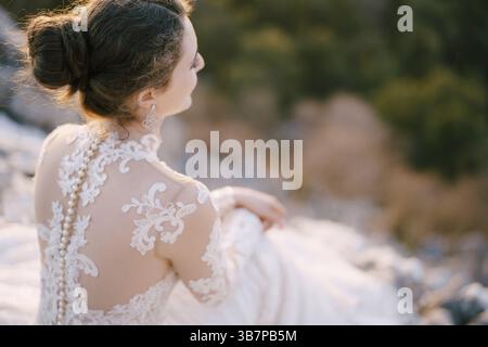 Braut in weißem Spitzenkleid sitzt auf den Steinen. Rückansicht. Stockfoto