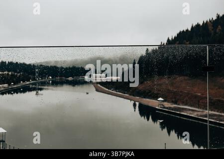 Malerischer Seeblick an einem regnerischen Tag durch ein Glasfenster mit Wassertropfen. Eine ruhige Landschaftsszene durch eine Glasbarriere, die von Regentropfen bedeckt ist Stockfoto