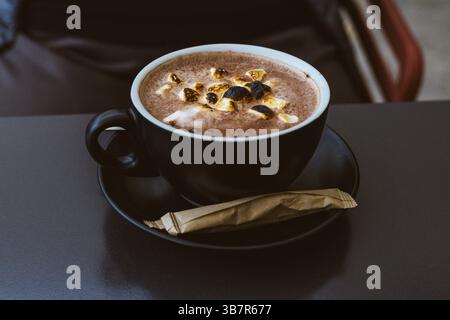 Eine dampfende schwarze Tasse heiße Schokolade mit Marshmallows auf einem Tisch. Eine gemütliche Szene mit einer warmen Tasse heißen Kakaos und gerösteten Marshmallows. Stockfoto