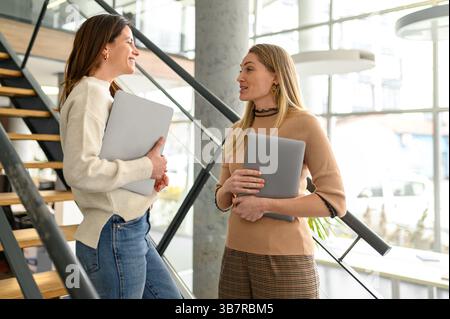 Weibliche Kollegen, die Computer halten und Projektdetails diskutieren, während sie in einem modernen Büro auf einer Treppe stehen Stockfoto