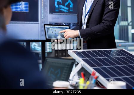 Investoren untersuchen die Oberfläche von Solarpaneelen mithilfe von Tablets und diskutieren die Effizienz der Konstruktion im Fabrikbüro. Geschäftsleute halten Gerätetreffen ab, um sich über technische Verbesserungen an Solarzellen zu unterhalten, Nahaufnahme Stockfoto