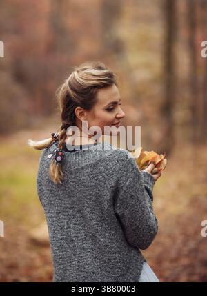 Attraktive Frau mit geflochtenen Zöpfen hält im Herbstwald gefallene Blätter in den Händen. Herbstkunstporträt Stockfoto