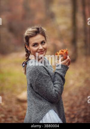 Attraktive Frau mit geflochtenen Zöpfen hält im Herbstwald gefallene Blätter in den Händen. Herbstkunstporträt Stockfoto
