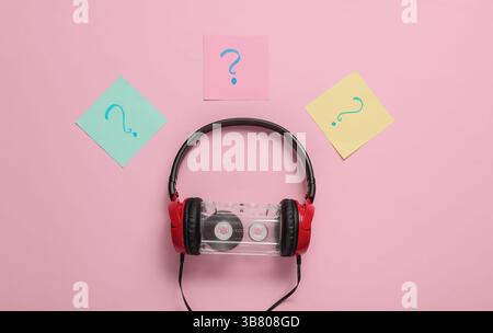 Kabelgebundene Kopfhörer mit Audiokassette und Fragezeichen auf Notizblättern auf rosa pastellfarbenem Hintergrund. Retro-Style, DJ. Blick von oben, minimalistischer Mus Stockfoto