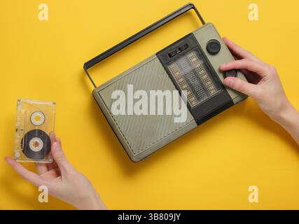 Weibliche Hand spielt Retro-Radioempfänger und hält die Audiokassette auf gelbem Hintergrund. Retro Mass Media, 70er Jahre Draufsicht Stockfoto