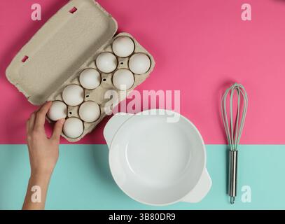 Der Prozess des Backens. Die weibliche Hand nimmt ein Ei aus einem Tablett auf rosa-blauem Hintergrund mit einer leeren Schüssel und einem Schneebesen. Draufsicht Stockfoto