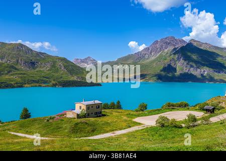 Der grüne Hügel führt hinunter zum einsamen Haus, dem Bergsee des Mont Cenis mit einem lebhaften türkisfarbenen Wasser und felsigen Bergen unter einem hellblauen Himmel. Stockfoto