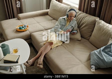 Ältere Frau genießt Musik mit Kopfhörern, während sie auf der Couch sitzt Stockfoto