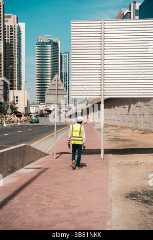 Dubai, VAE - 30. April 2026: Asiatischer Migrant Worker auf den Straßen von Dubai. Hochwertiges fotooffenes Bild eines asiatischen Arbeitsmigranten, der auf einer Straße in Dubai, Vereinigte Arabische Emirate, spaziert. In Arbeitskleidung unter der intensiven Sonne des Nahen Ostens verkleidet, repräsentiert der Mann die hart arbeitenden Einwanderer, die die rasante Stadtentwicklung der Stadt unterstützen. Das Foto zeigt Themen wie Globalisierung, Arbeitsmigration und Alltag in einer modernen Metropole. Stockfoto