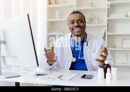 Schwarzer Männlicher Arzt Sitzt Am Schreibtisch Mit Computer Und Spricht Vor Der Kamera Stockfoto