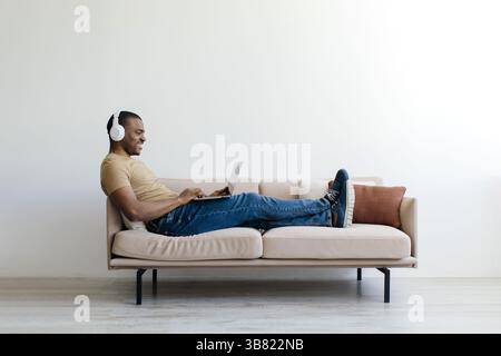 Junger Schwarzer Mann, Der Drahtlose Kopfhörer Trägt Und Sich Auf Der Couch Mit Laptop Entspannt Stockfoto