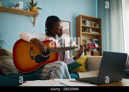 Afroamerikanerin genießt Gitarrenübungen im Wohnzimmer, während sie auf einer bequemen Couch sitzt. Die Umgebung umfasst Laptop und Bookshe Stockfoto