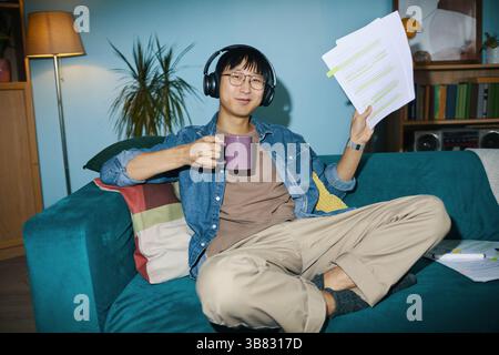 Porträt eines jungen Mannes, der Kaffee genießt, während er von zu Hause aus arbeitet, Dokumente hält und Kopfhörer trägt. Sitzen auf blaublauem Sofa in einem organisierten Wohn r Stockfoto