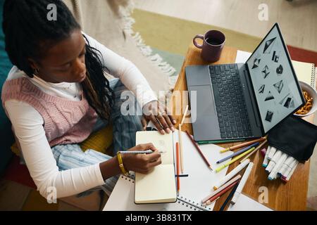 Frau, die in einem Notizbuch schreibt, umgeben von einer Vielzahl von Untersuchungsmaterialien und einem offenen Laptop auf einem Holztisch, zeigt einen produktiven Arbeitsplatz Stockfoto